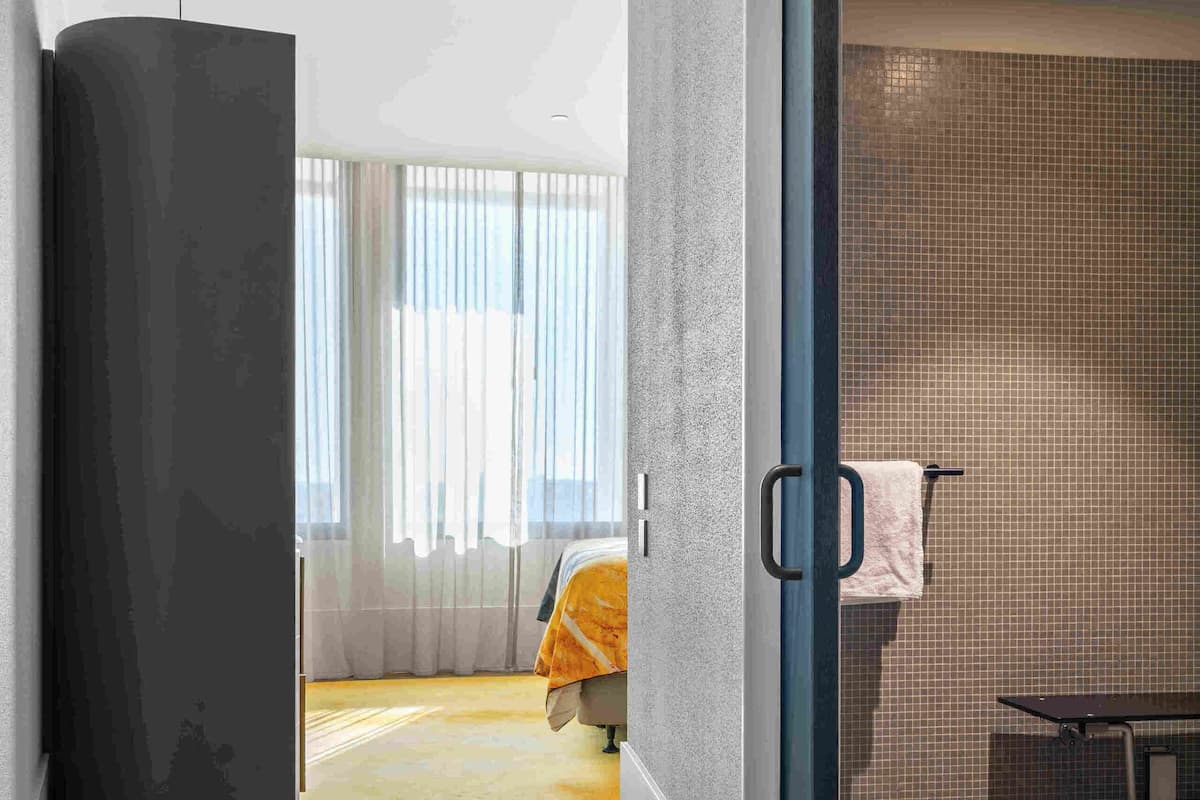 Camera d'albergo moderna con pavimento in parquet a spina di pesce, una grande finestra con tende leggere e bagno in vista con rivestimento in piastrelle.
