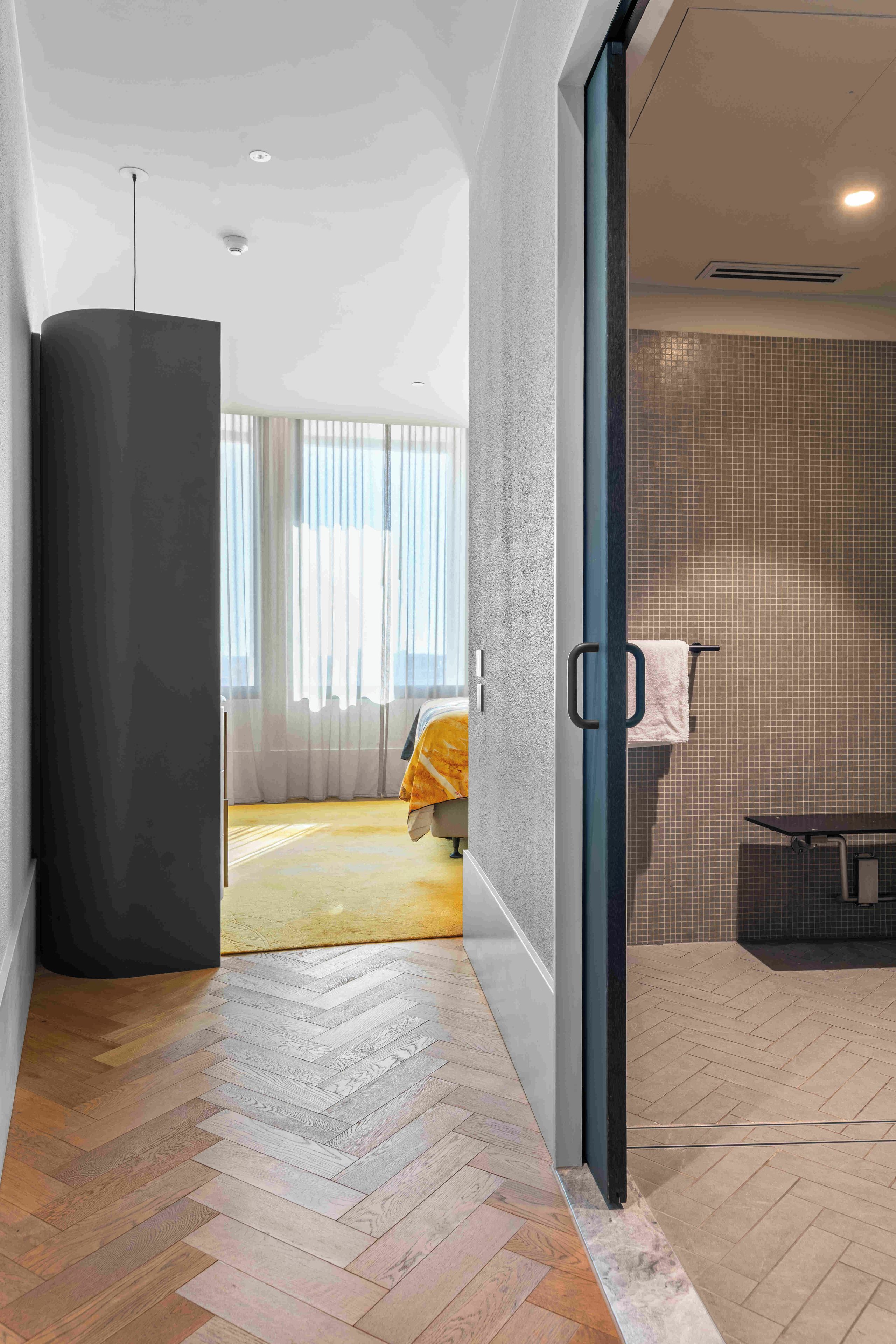 A modern hotel room with a herringbone wood floor, a large window with sheer curtains, and a visible bathroom with a tiled shower wall.
