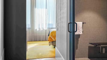 A modern hotel room with a herringbone wood floor, a large window with sheer curtains, and a visible bathroom with a tiled shower wall.
