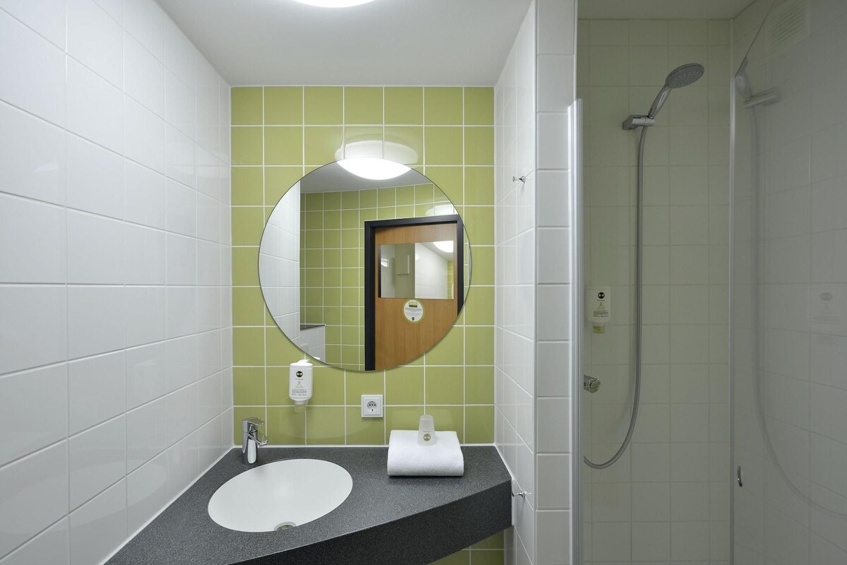 Een badkamer met een wit wastafel, een ronde spiegel, een douche en groene tegels.