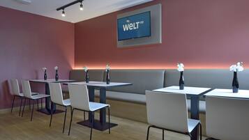 Un comedor moderno con mesas y sillas, una pantalla grande mostrando 'welt HD', y un banco a lo largo de la pared.