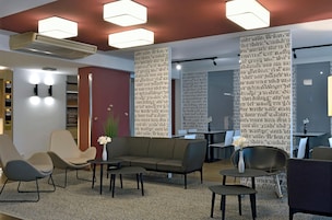 Un área de estar moderna con sofá, sillones y mesas. Hay unidades de aire acondicionado y una pared decorativa con texto.