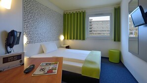 Habitación de hotel con cama, escritorio con control remoto y revista, otomana verde, televisor montado en la pared y ventana con cortinas.