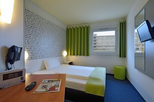 A hotel room with a bed, a desk with a remote control and a magazine, a green ottoman, a TV mounted on the wall, and a window with curtains.