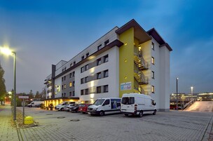 Un hotel a più piani con una parete gialla decorativa, un'area di parcheggio con diversi veicoli e una scala che conduce a un livello superiore.