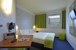 A hotel room with a bed, a desk with a remote control, a magazine, a wall-mounted phone, and a TV on the wall. - B&B Hotel München Messe Dornach (Aschheim)
