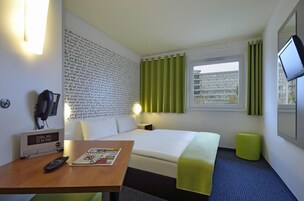 Una camera d'albergo con un letto, una scrivania con un telecomando, una rivista, un telefono fissato al muro e una TV a parete.