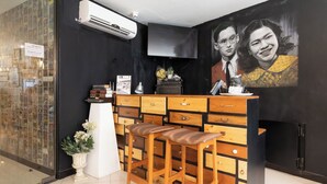 A reception area with a black wall, a mounted TV, a wooden counter with drawers, and two wooden stools. - The Secret Service Hotel (Bangkok)