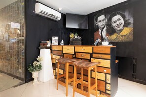 A reception area with a black wall, a mounted TV, a wooden counter with drawers, and two wooden stools. - The Secret Service Hotel (Bangkok)
