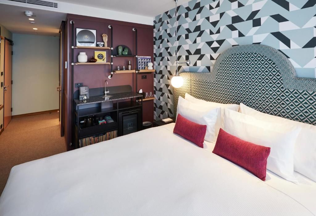 Habitación de hotel con una cama grande, cabecera con diseño geométrico, estante empotrado con varios objetos y pared con diseño de papel tapiz geométrico.