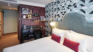 Habitación de hotel con una cama grande, cabecera con diseño geométrico, estante empotrado con varios objetos y pared con diseño de papel tapiz geométrico.