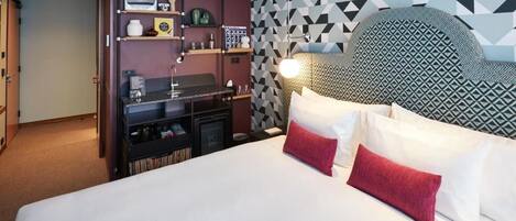 A hotel room with a large bed, a headboard with a geometric pattern, a built-in shelf unit with various items, and a wall with a geometric wallpaper design.