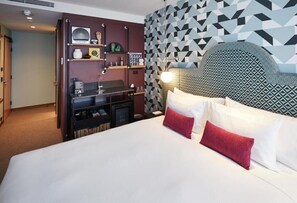Une chambre d’hôtel dotée d’un grand lit, d’une tête de lit ornée d’un motif géométrique, d’une bibliothèque intégrée contenant divers objets et d’un mur recouvert d’un papier peint à motifs géométriques.