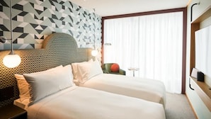 Habitación de hotel con dos camas, cabecera de diseño geométrico, una mesita de noche con lámpara y vistas al exterior a través de cortinas translúcidas.