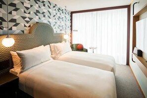 Une chambre d’hôtel avec deux lits, une tête de lit à motifs géométriques, une petite table avec une lampe et une vue sur l’extérieur à travers des rideaux transparents.
