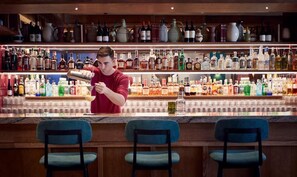 Um balcão de bar com estantes cheias de garrafas de bebidas e um barman preparando uma bebida.