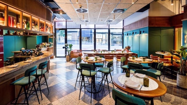 Un restaurante con zona de bar, varias mesas y sillas, y amplios ventanales que dan a la calle.