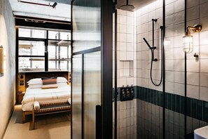 Ein modernes Badezimmer mit Dusche, Toilette und Waschbecken. Im angrenzenden Zimmer befindet sich ein Bett mit einem Fenster.