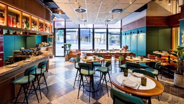 Een restaurant met een bar, meerdere tafels en stoelen, en grote ramen met uitzicht op de straat.