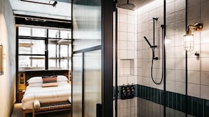 Een moderne badkamer met een douche, toilet en wastafel. In de aangrenzende kamer bevindt zich een bed met een raam.