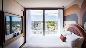 Habitación de hotel con una cama grande, un televisor de pantalla plana montado en la pared y vistas de la ciudad a través de un ventanal amplio.