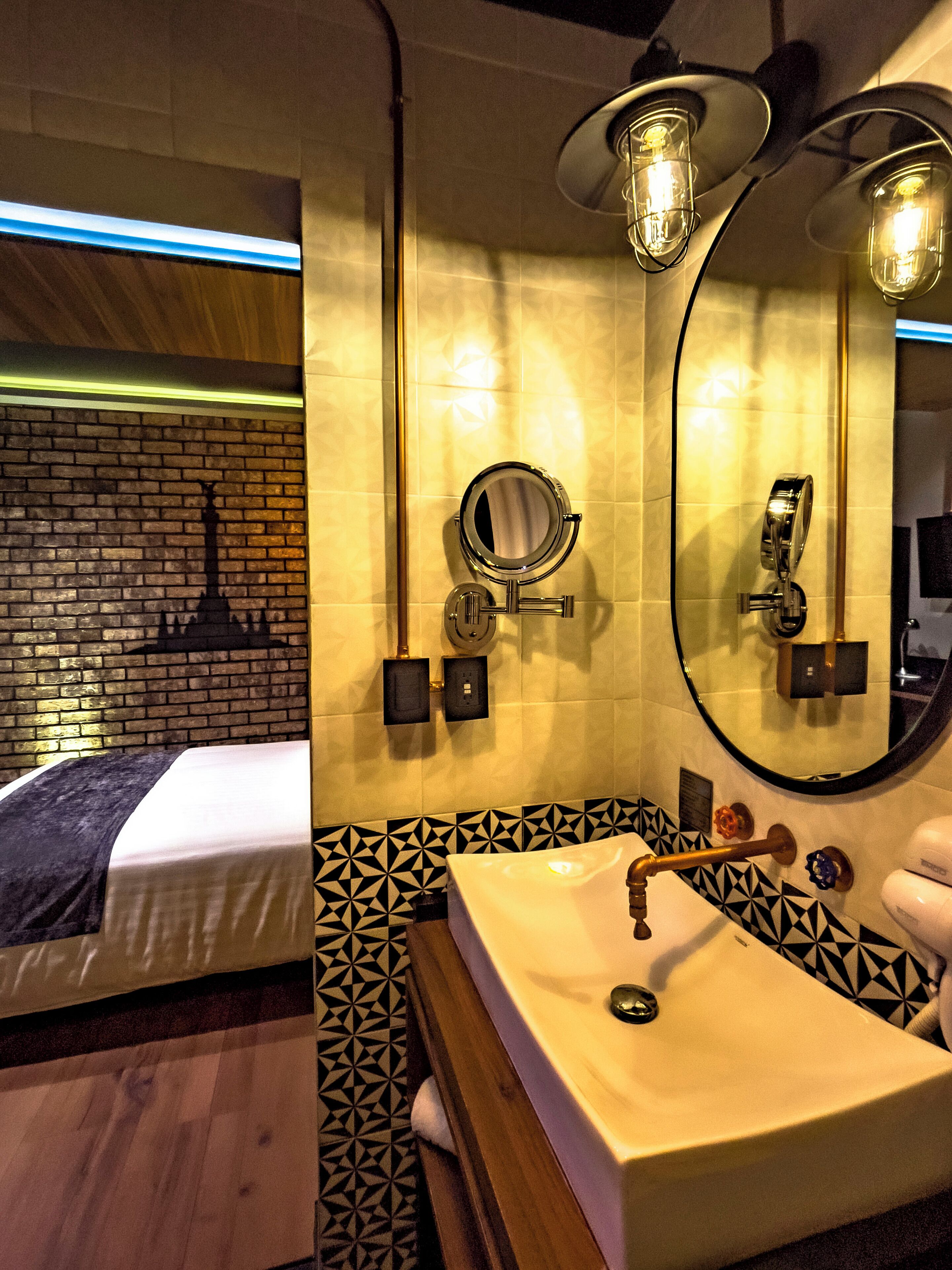 Ein Hotelbad mit Waschbecken, Spiegel und Handtuchhalter. Die Wand ist mit einem Muster versehen und im Hintergrund steht ein Bett.