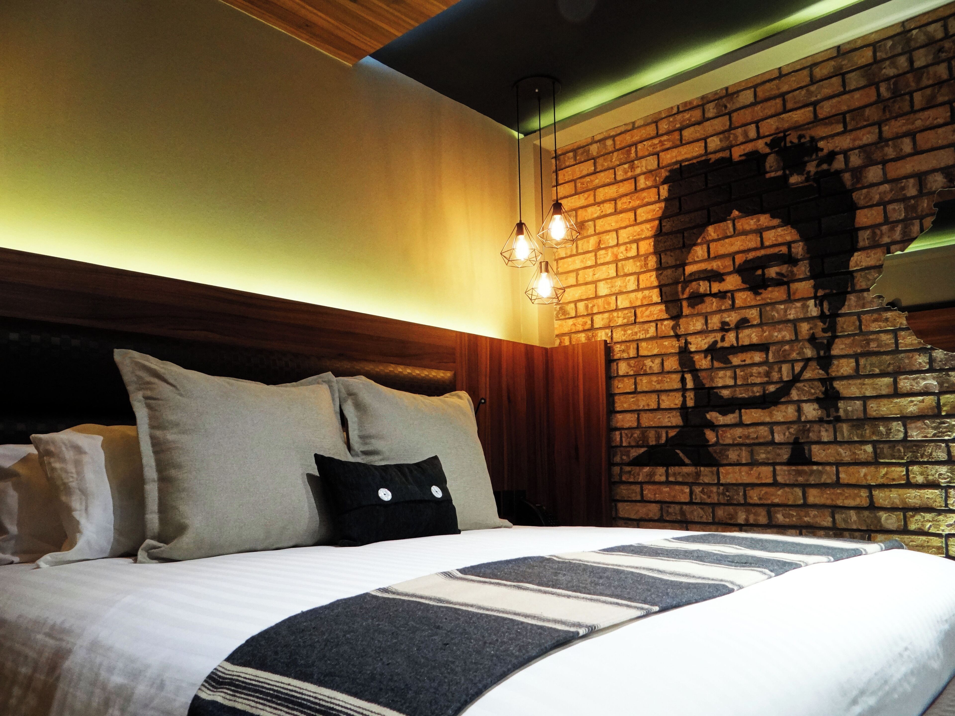 Ein Schlafzimmer mit Wandgemälde an einer Ziegelwand, einem Bett mit gestreifter Bettwäsche und hängenden Pendelleuchten.