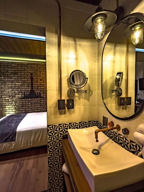 A hotel bathroom with a sink, mirror, and towel rack. There is a bed with a headboard in the background. - Boutique Hotel Cadillac (Mexico City)