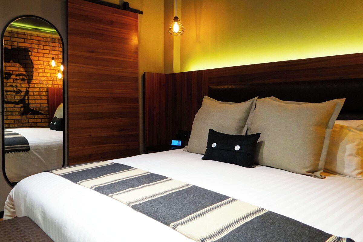 A modern hotel room with a large bed, wooden headboard, and a mirror reflecting a brick wall.