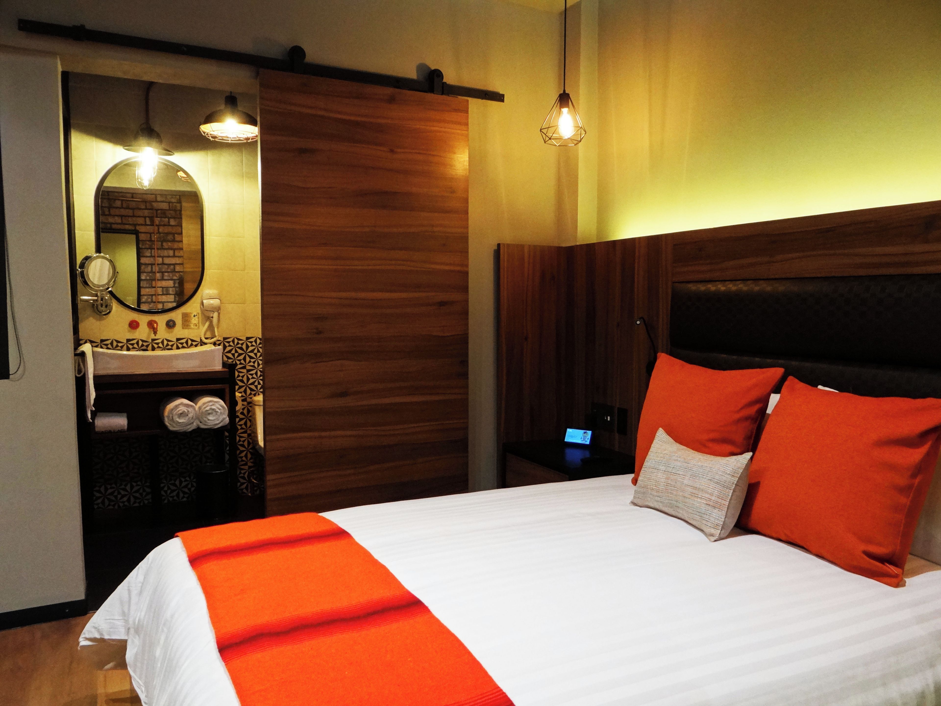 A hotel room with a bed, a wooden headboard, a nightstand, a mirror, a sink, and a towel rack.