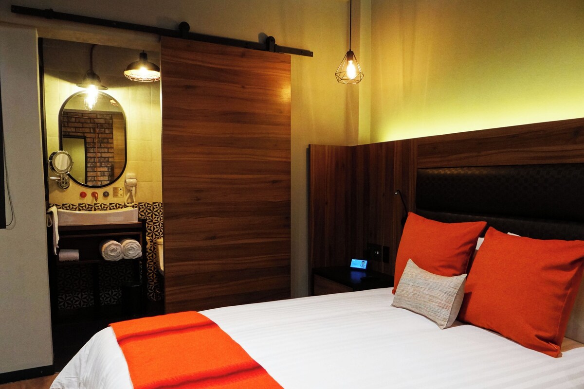 A hotel room with a bed, a wooden headboard, a nightstand, a mirror, a sink, and a towel rack.