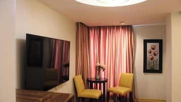 A hotel room with a flat-screen TV, two yellow chairs, a small round table with a vase of flowers, and a large window with striped curtains.