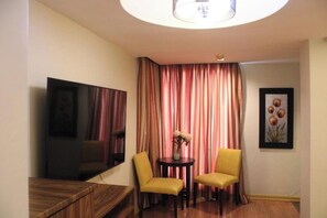 Quarto de hotel com televisão de tela plana, duas cadeiras amarelas, uma mesa redonda pequena com um vaso de flores e uma janela grande com cortinas listradas.