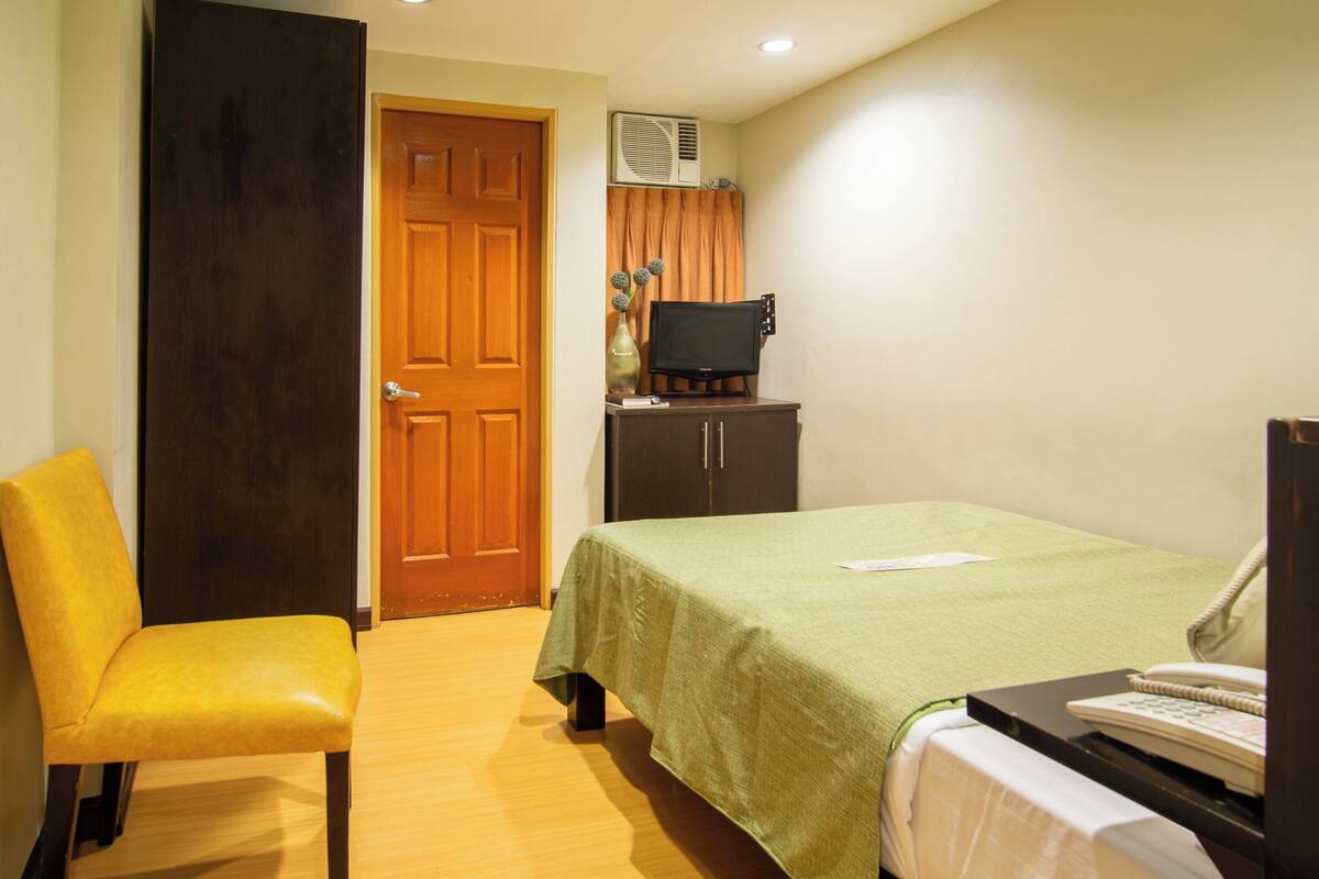 Una habitación de hotel con cama, una silla, escritorio con teléfono, televisor y armario.
