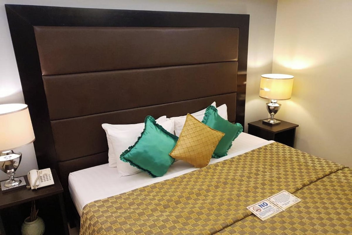 A hotel room with a large bed, multiple pillows, a bedside lamp, a phone, and a sign on the bed.