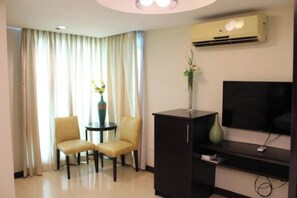 Quarto de hotel com televisão de tela plana, um armário de madeira escura, duas cadeiras, uma mesa redonda pequena e uma janela com cortinas.