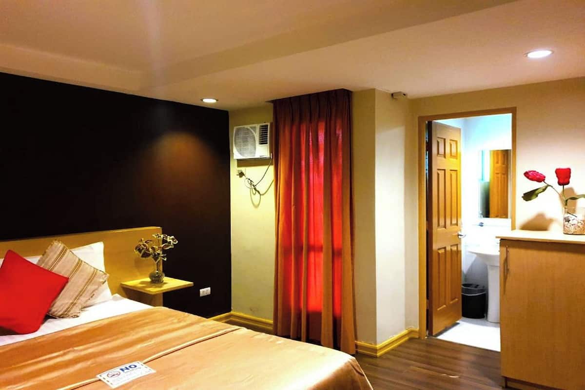 Habitación de hotel con cama, mesita de noche con un florero, cortina roja y puerta de madera que da a un baño.