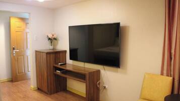 Een woonkamer met een flatscreen televisie aan de muur, een houten tv-meubel, een gele bank en een deur naar een andere kamer.