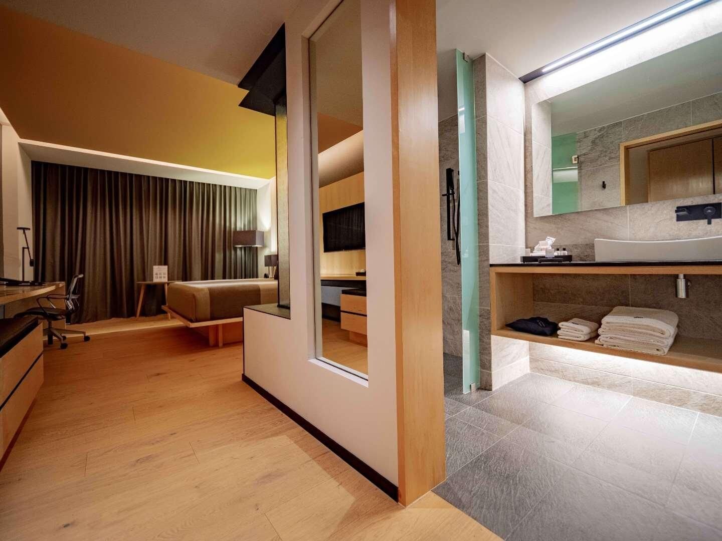 A modern hotel room with a sliding door leading to a balcony, a separate living area with a sofa and a desk, and a bathroom with a large mirror and a vanity.