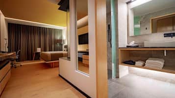 A modern hotel room with a sliding door leading to a balcony, a separate living area with a sofa and a desk, and a bathroom with a large mirror and a vanity.