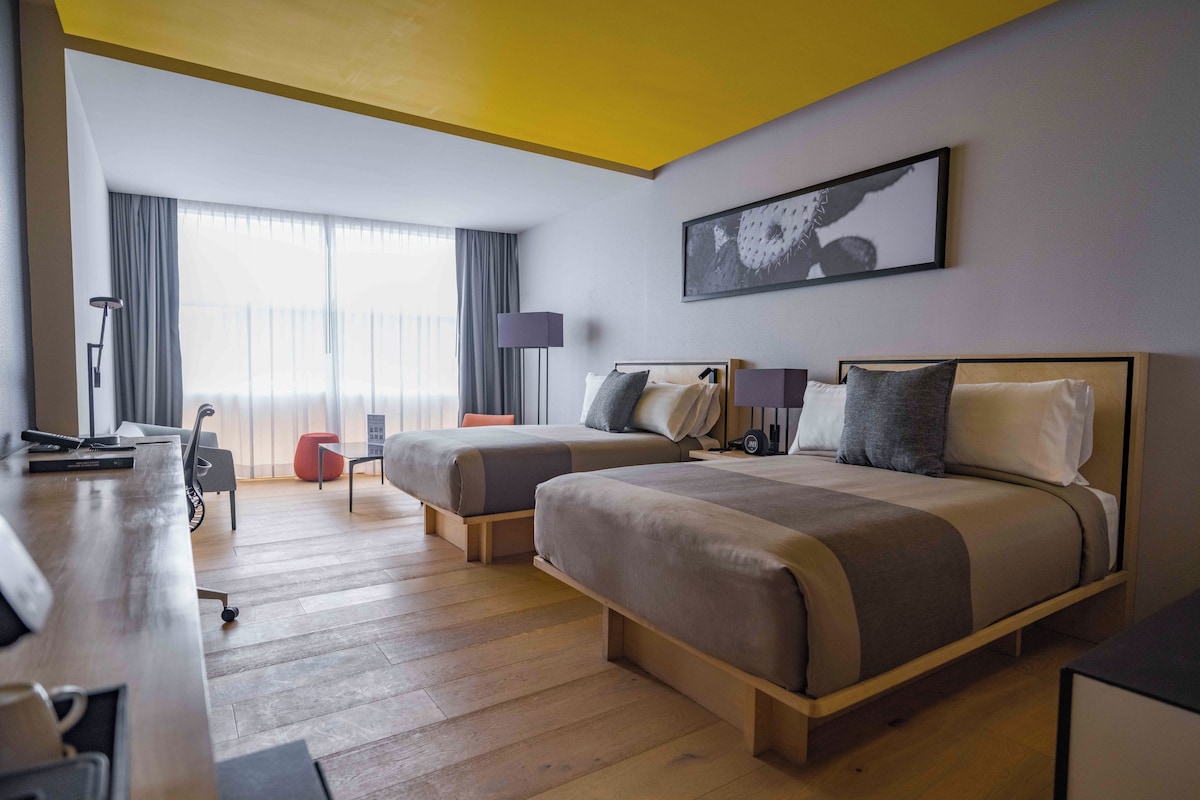 A hotel room with two beds, a desk, a lamp, and a large window with curtains.
