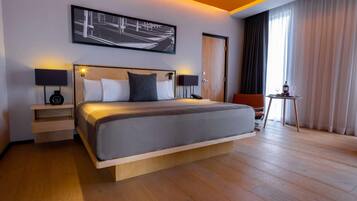 A modern hotel room with a large bed, bedside tables with lamps, a small table with wine glasses, and a view of the outdoors through large windows.