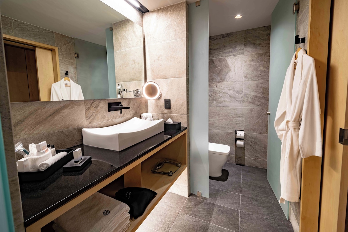 A modern bathroom with a curved sink, a mirror with a light, a toilet, and a robe hanging on a door.