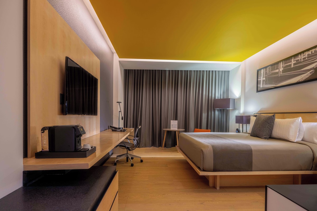 A hotel room with a bed, a desk with a computer, a TV, and a window with curtains.