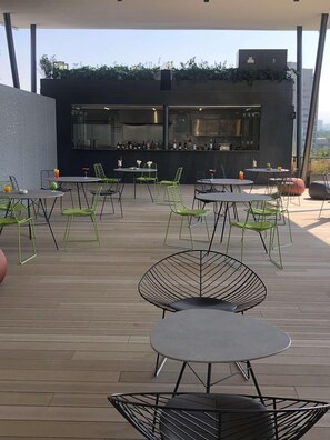 Un patio exterior con varias mesas y sillas, un área de bar con botellas y vistas al horizonte de la ciudad.