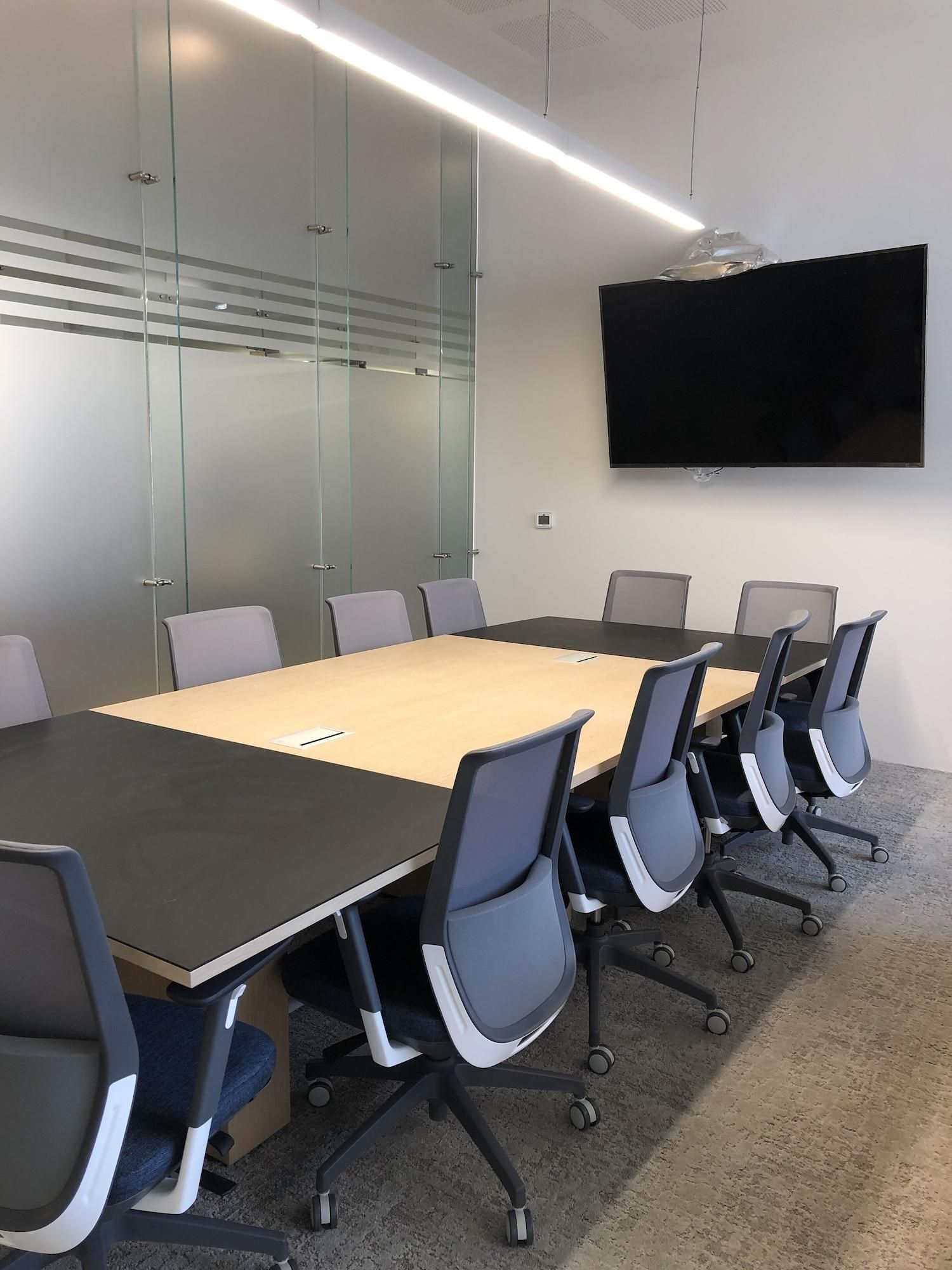 A conference room with a long table, six chairs, a flat-screen TV, and glass walls.