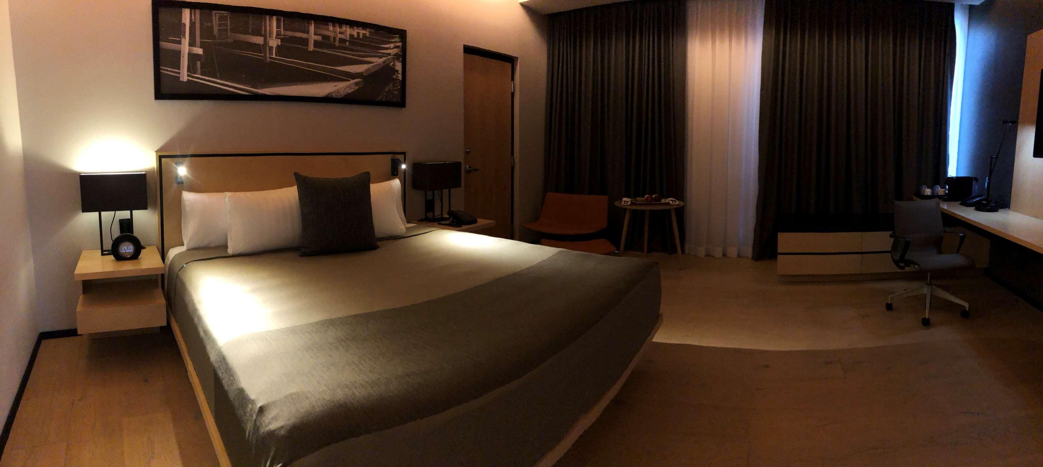 A hotel room with a large bed, a desk, a chair, a small table, and a nightstand with a lamp.