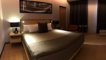 A hotel room with a large bed, a desk, a chair, a small table, and a nightstand with a lamp.