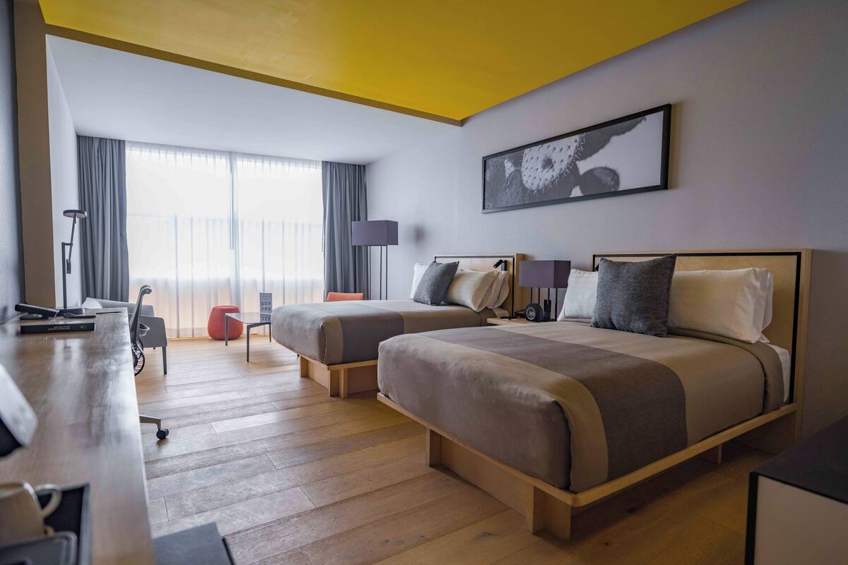 Une chambre d’hôtel avec deux lits, un bureau, une lampe et une grande fenêtre avec des rideaux.