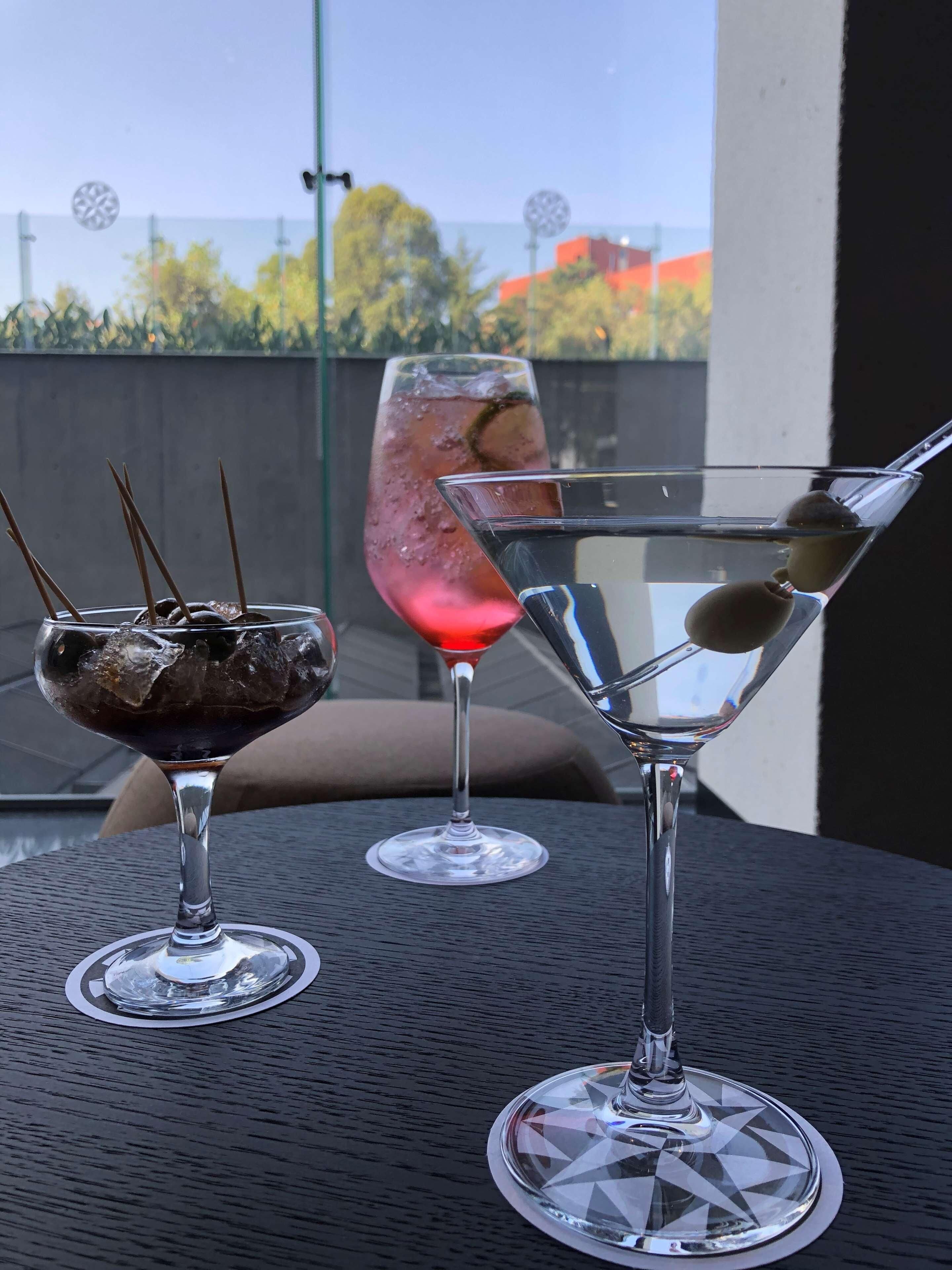 Três copos com bebidas sobre uma mesa, com vista para árvores e edifícios ao fundo.